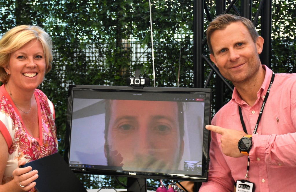Woman and man standing, smiling, next to a monitor showing the face of a team member.